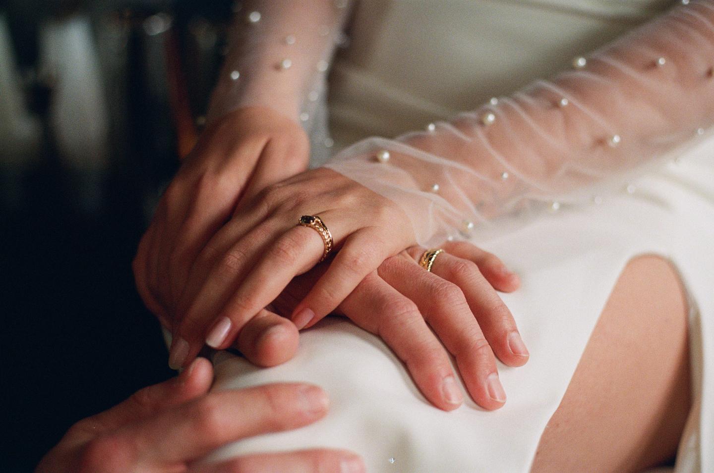 A couple's hands are intertwined, with the woman wearing a sheer, pearl-embellished sleeve and a ring with a dark stone, and the man wearing a gold band.