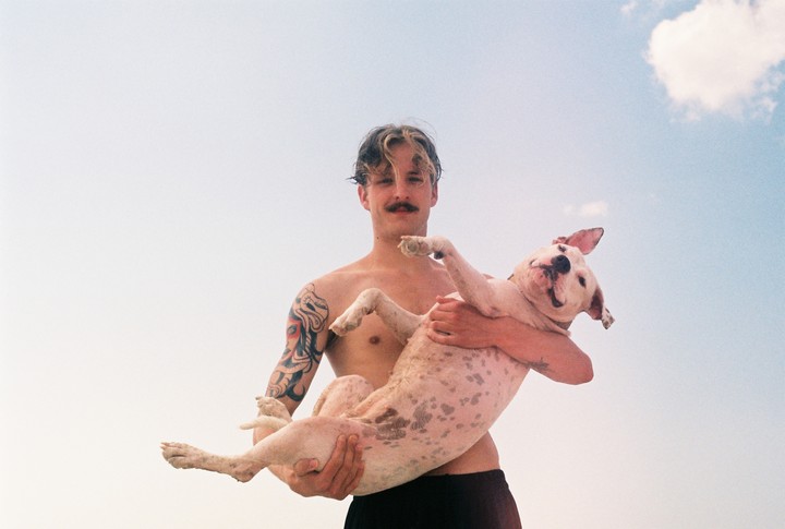 A man with a mustache and tattoos holds a dog against a clear sky with a single cloud.