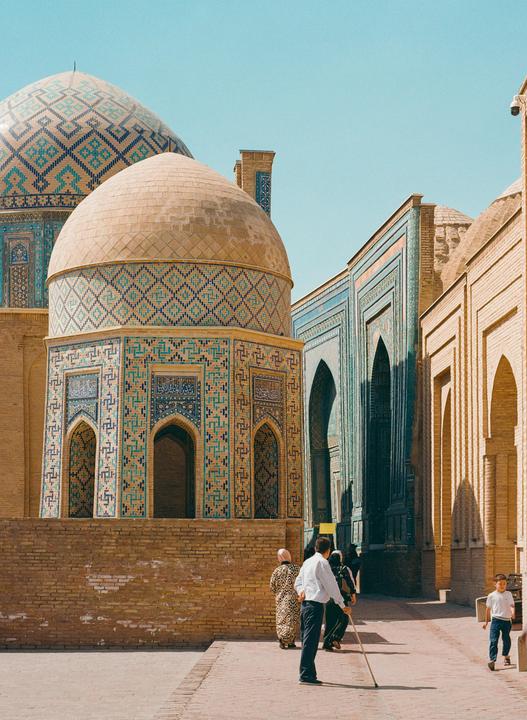 People walk past ornate tiled buildings with domes under a clear sky.