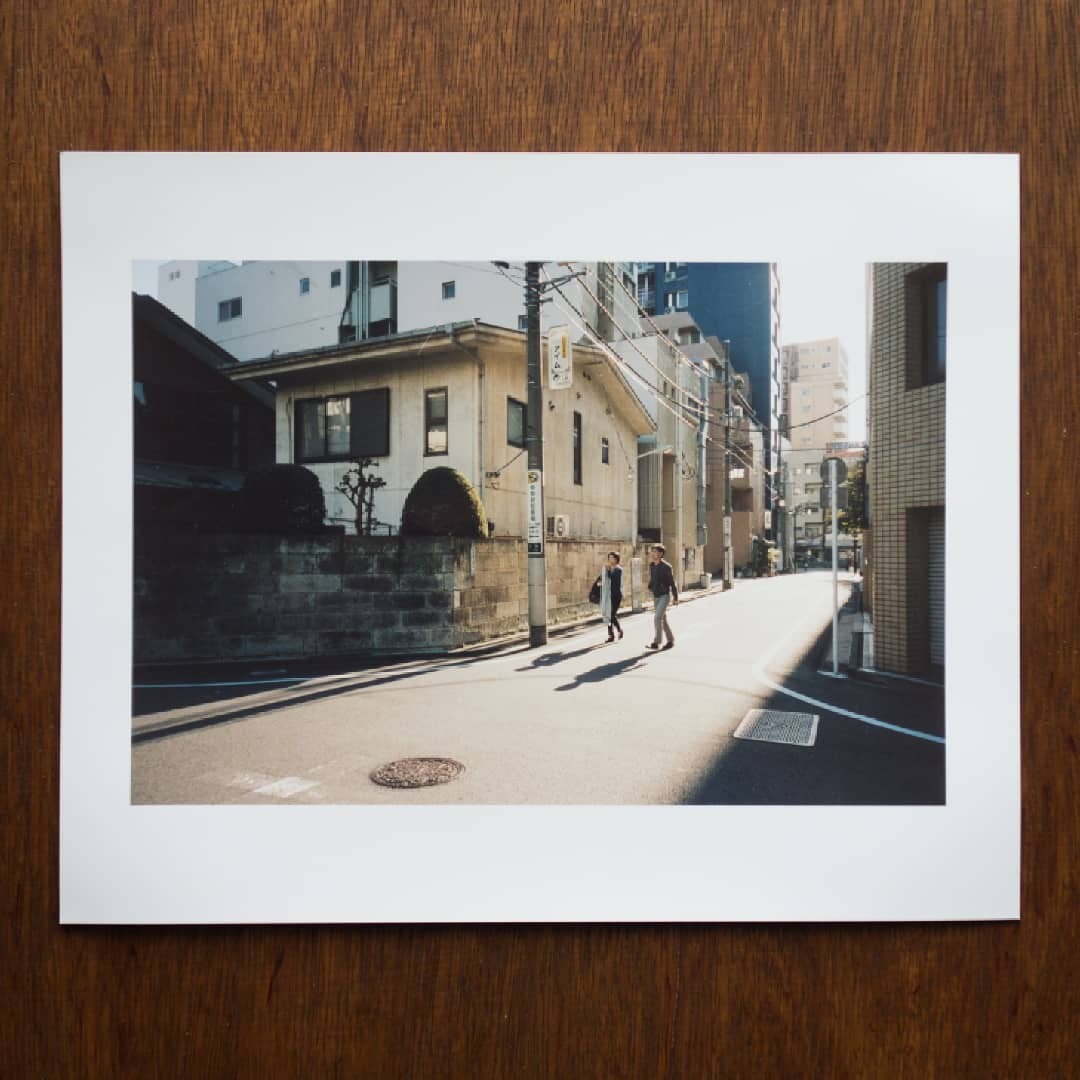 Two people walk across a sunlit street in a city with buildings and power lines.