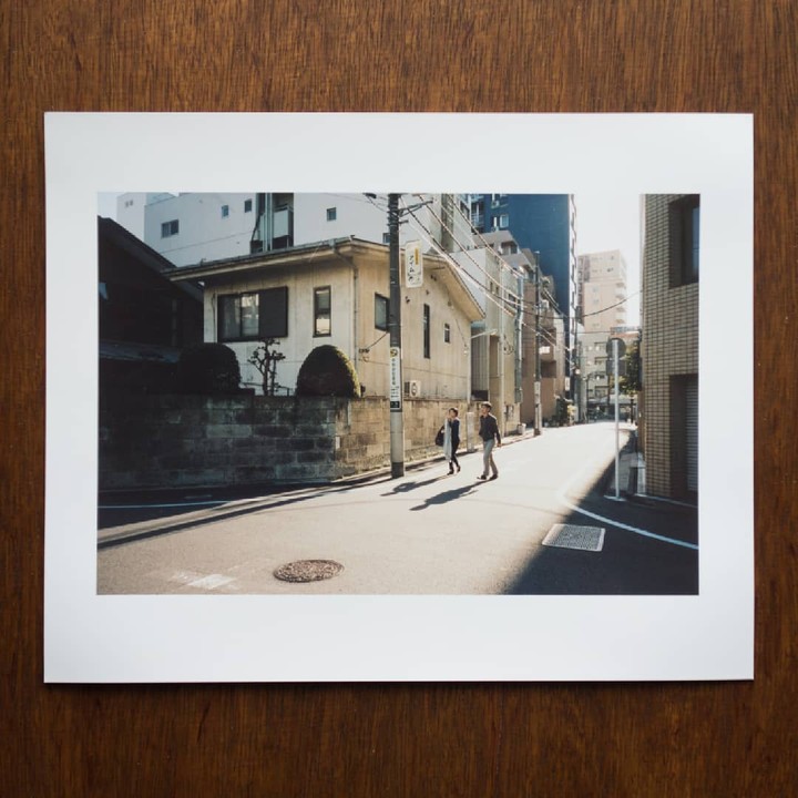 Two people walk across a sunlit street in a city with buildings and power lines.