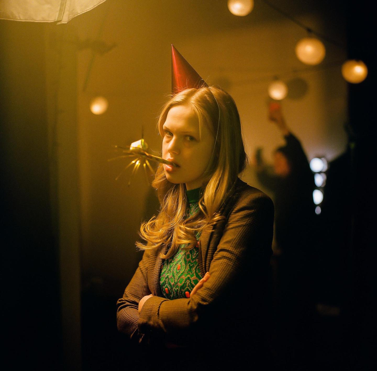 A woman wearing a party hat and holding a party blower stands with her arms crossed in a dimly lit room.