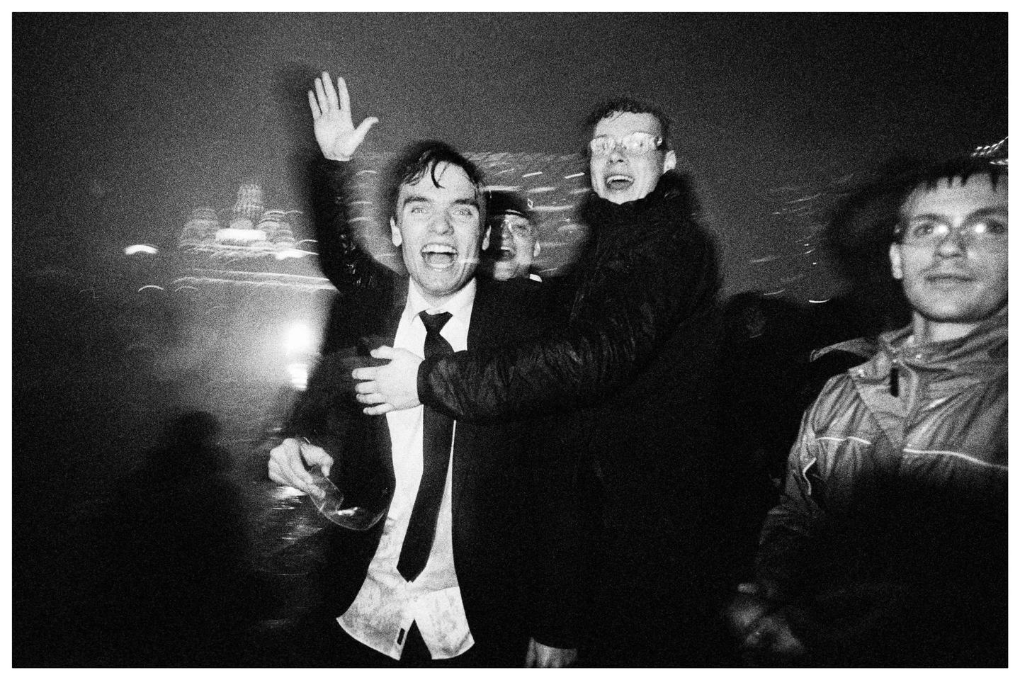 A man in a suit with his arm raised and mouth open stands next to another man in a dark jacket, with two other men visible behind them.