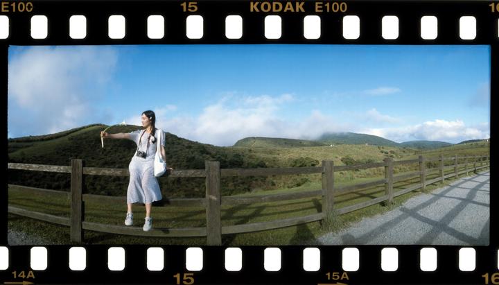 A woman sits on a wooden fence in a panoramic landscape with rolling green hills and a blue sky.