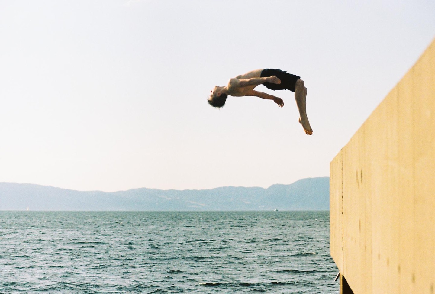 A man performs a backflip off a wooden pier over a body of water with mountains in the distance.