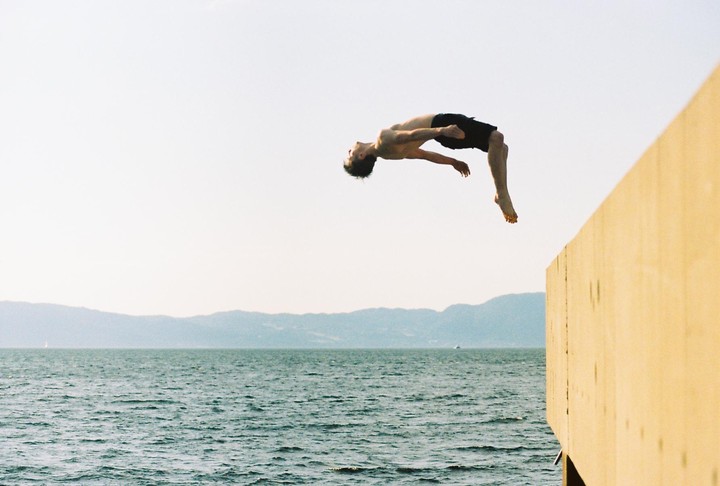 A man performs a backflip off a wooden pier over a body of water with mountains in the distance.