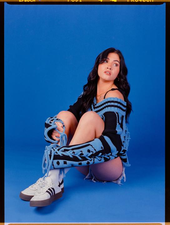 A woman wearing a blue and black sweater and white sneakers sits on a blue surface.