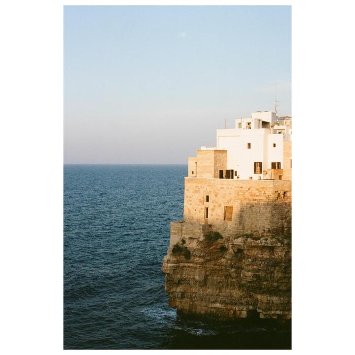 A white building with brown shutters sits atop a cliff overlooking the ocean.