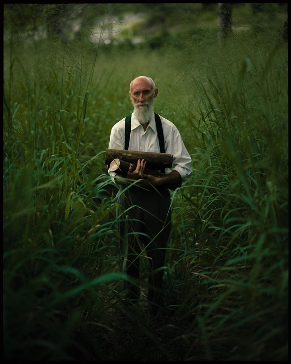 A man with a long white beard stands in tall grass, holding logs.