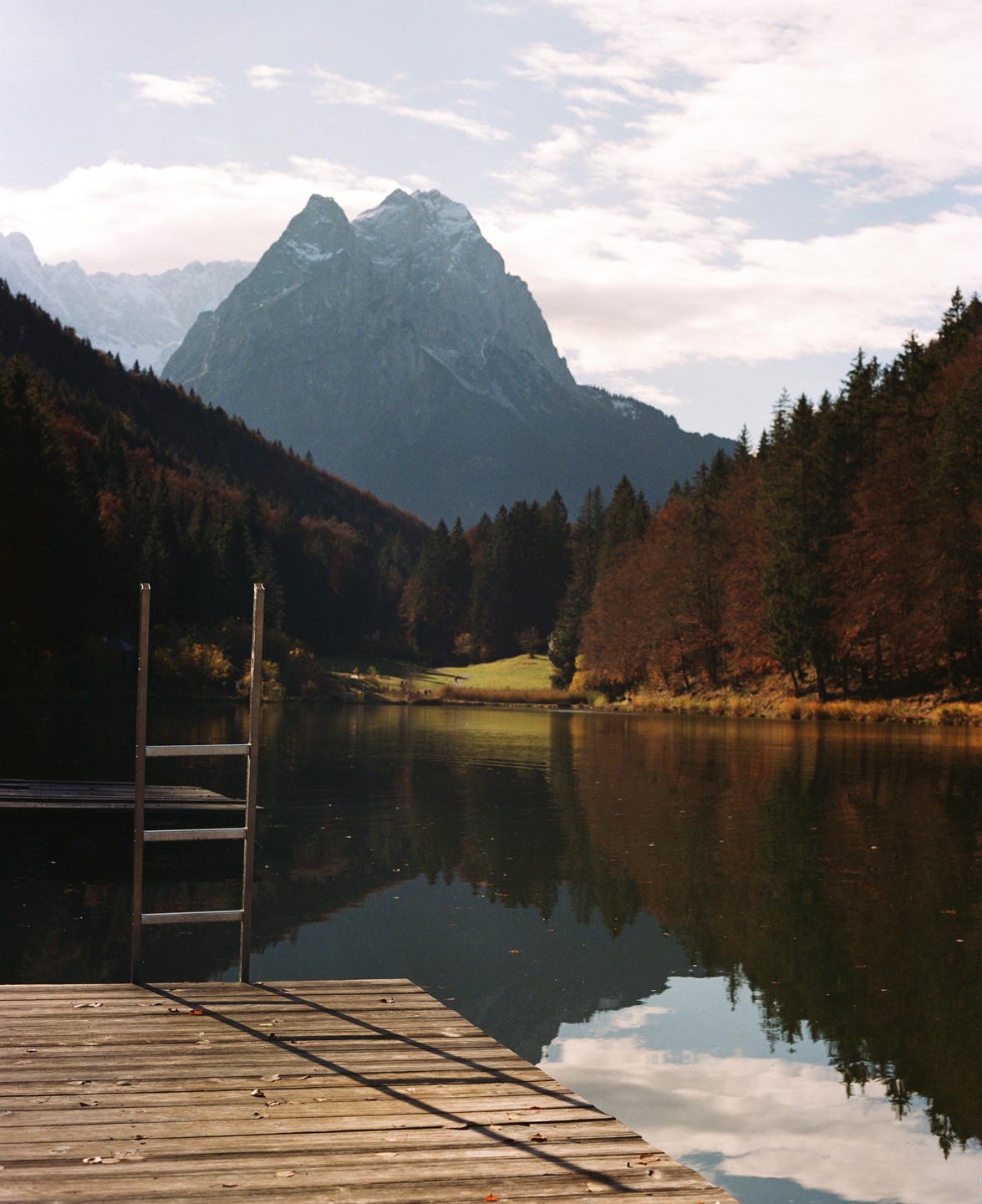 A wooden dock with a ladder extends into a calm lake, reflecting mountains and autumn trees under a cloudy sky.