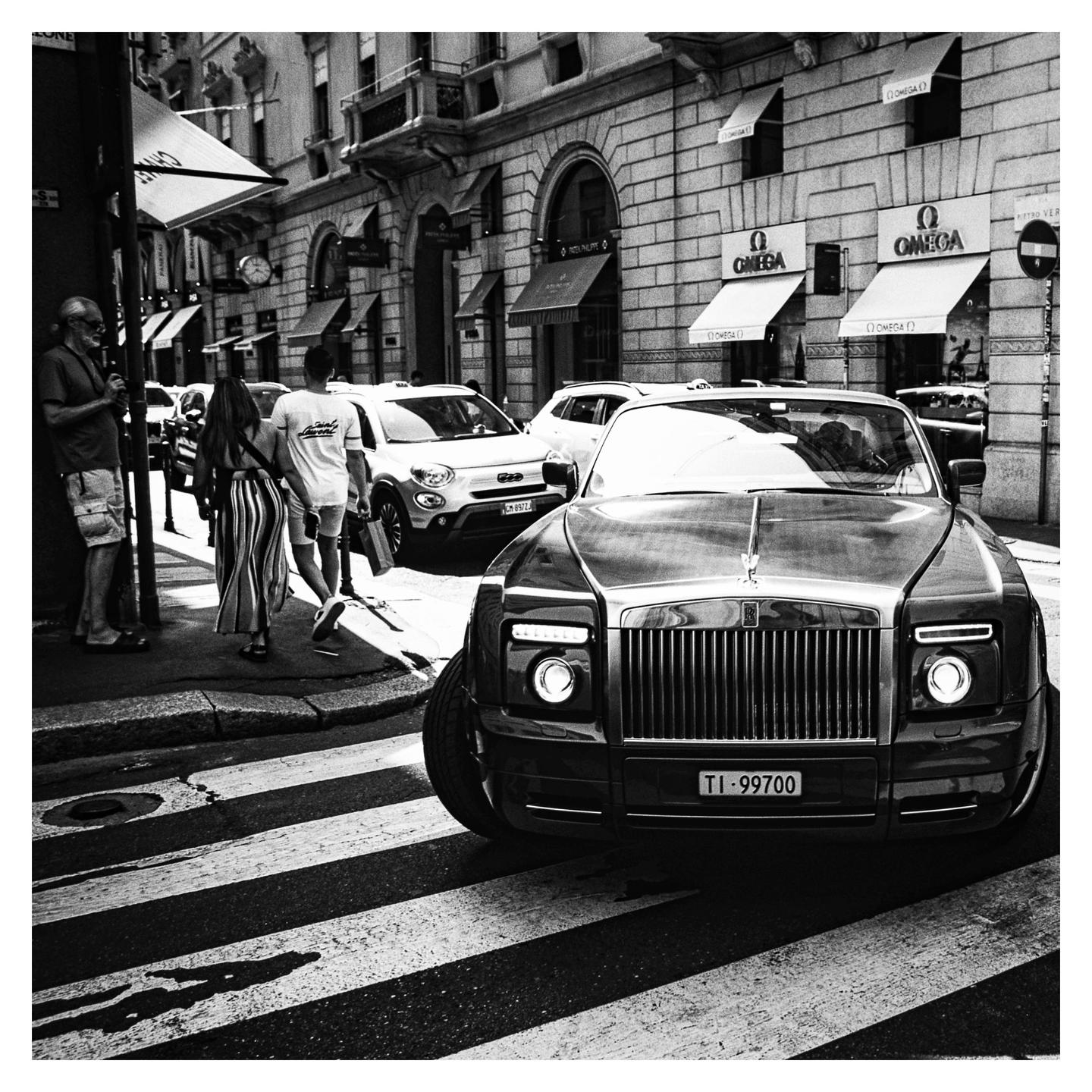 A luxury car is stopped at a crosswalk on a city street with people walking on the sidewalk.
