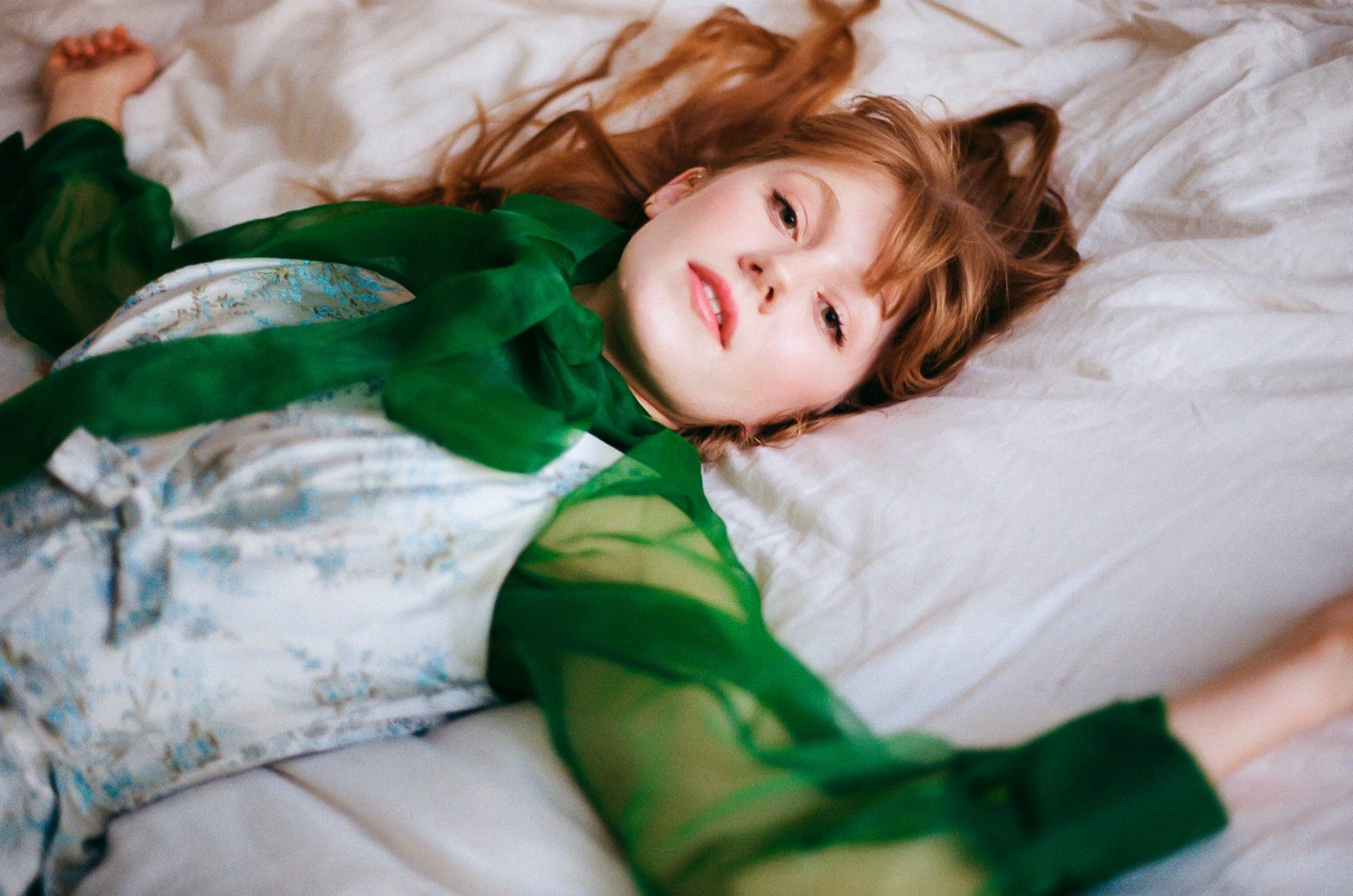 A woman with long red hair and a green sheer top lies on a white bed.