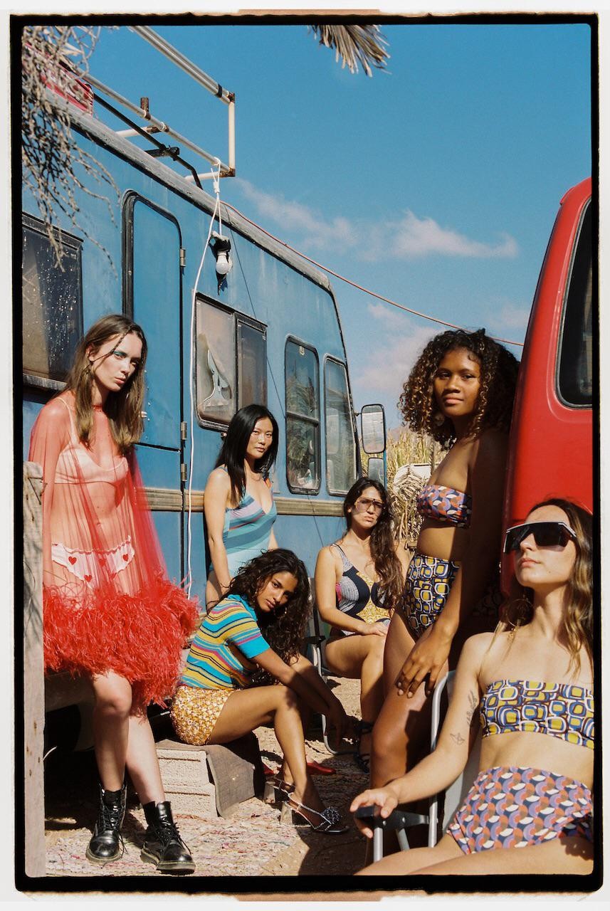 Six women pose in front of a blue camper van and a red vehicle under a clear sky.