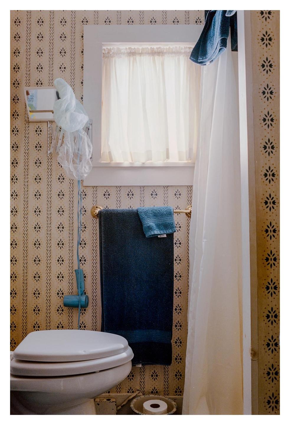 A toilet is visible in a bathroom with patterned wallpaper, a window with a sheer curtain, and blue towels hanging on a rack.