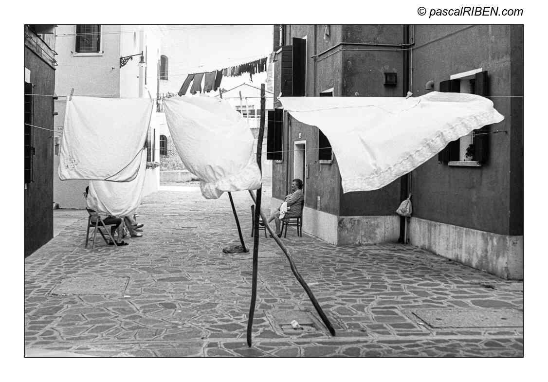 Two women sit on chairs in a narrow alleyway with laundry hanging overhead and on lines in the distance.