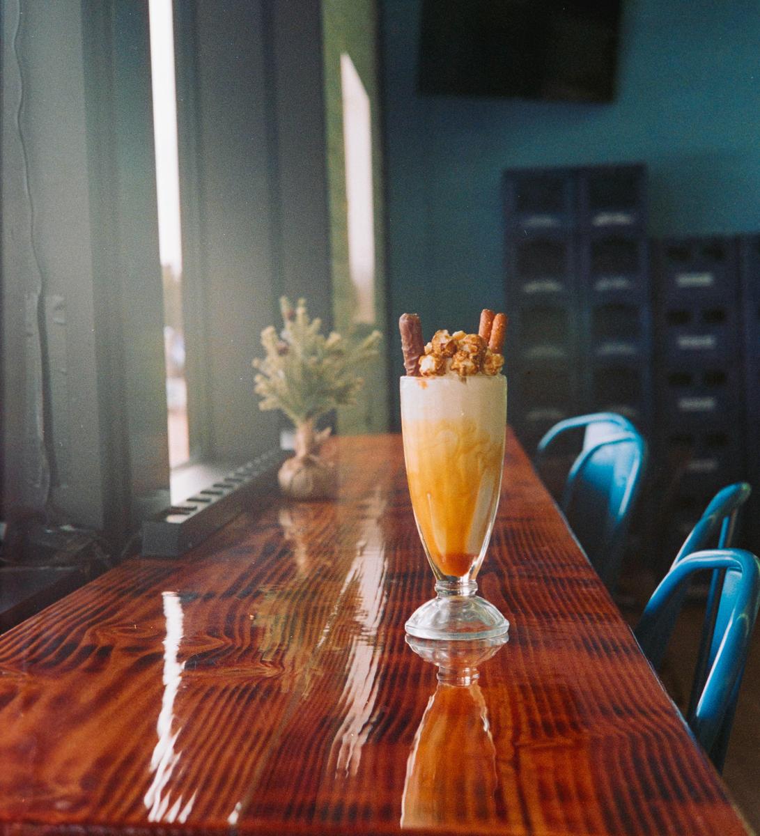 A tall glass of ice cream with toppings sits on a glossy wooden counter next to a window.