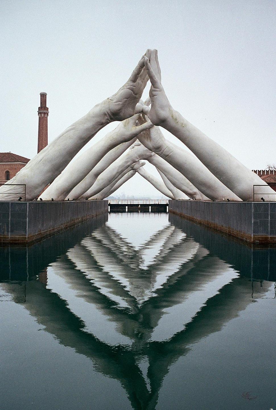 A sculpture of giant hands forming arches over a canal is reflected in the water.