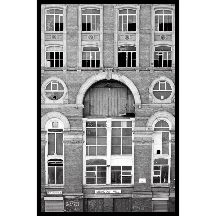 The facade of Meadow Mill is shown with many windows, some broken, and an arched entrance.