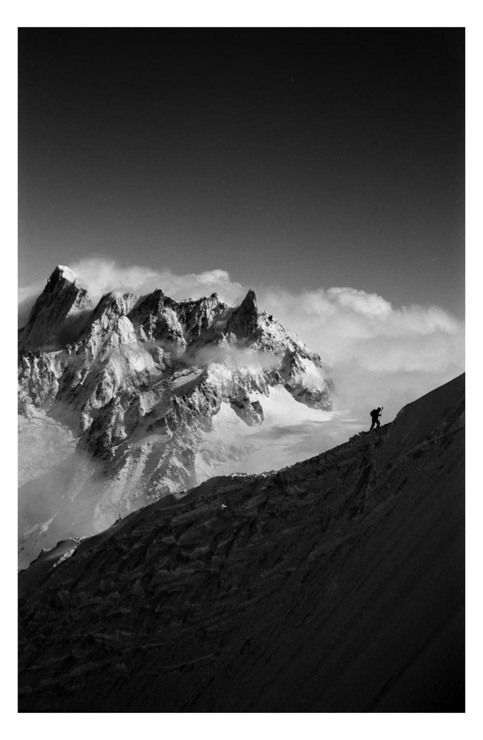 A lone climber ascends a steep, rocky slope with snow-capped mountains and clouds in the background.