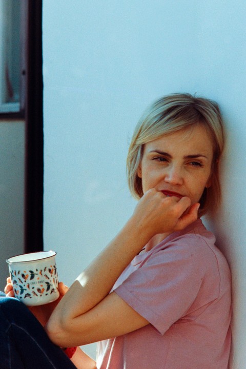 A woman with short blonde hair leans against a light blue wall, holding a patterned mug.
