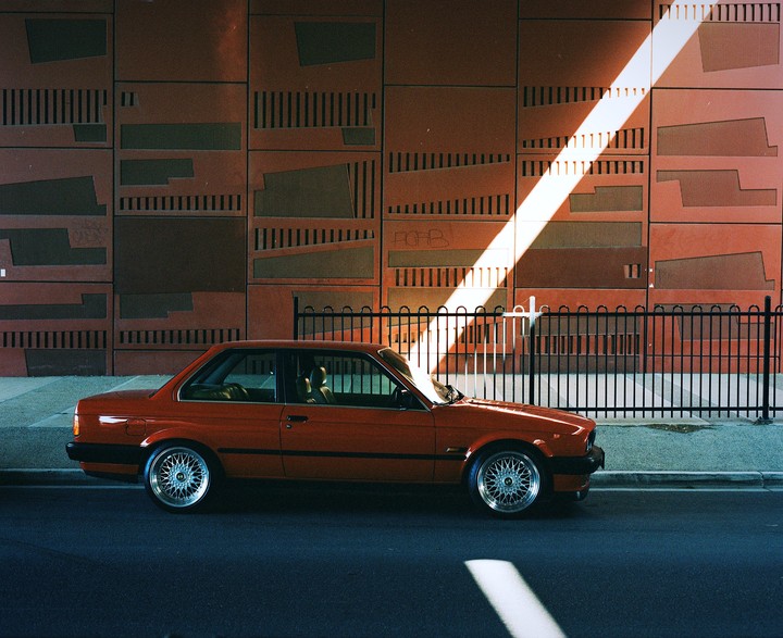 A red car is parked on a street next to a fence and a building with a diagonal beam of light shining on it.