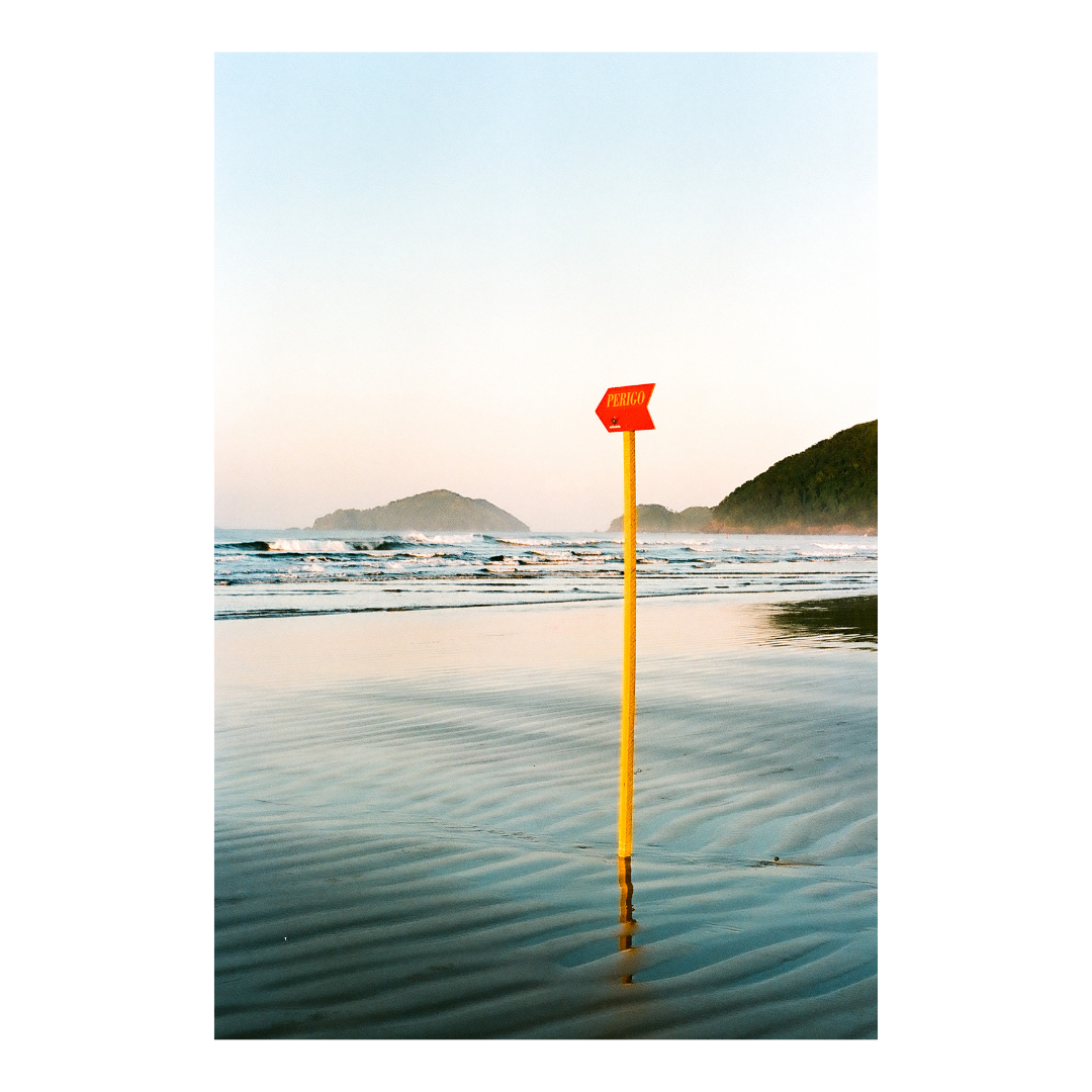 A red flag with the word "PERIGO" on a yellow pole stands in the wet sand of a beach with waves and distant hills under a pale sky.