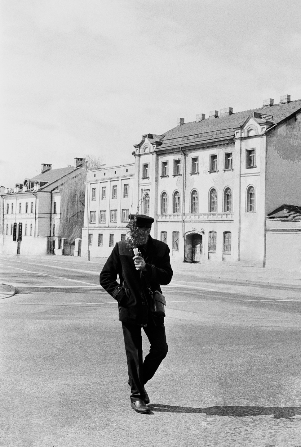 A man in a dark coat and cap walks across a street holding a flower, with buildings in the background.