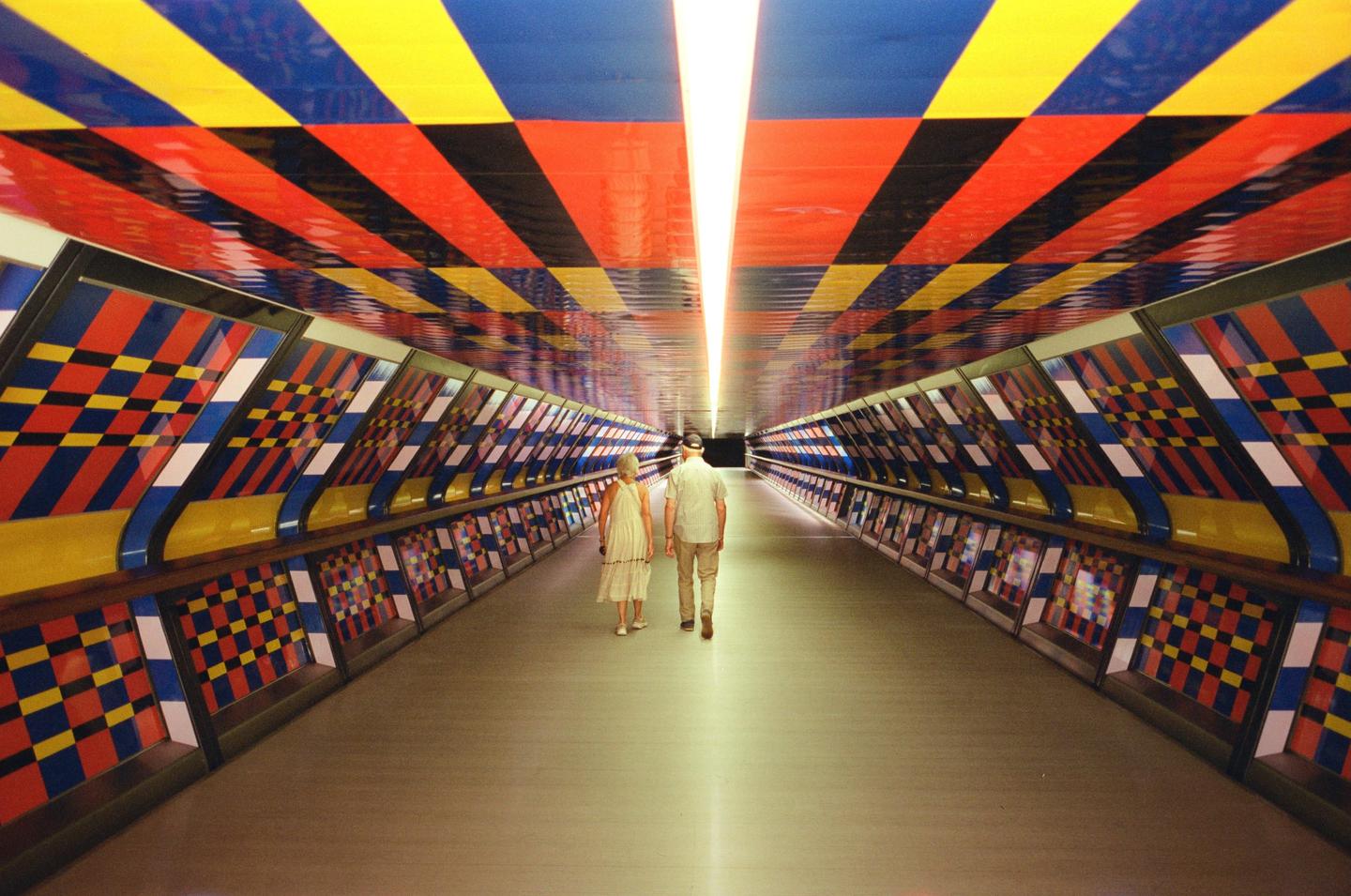 A couple walks away down a long, brightly lit tunnel with colorful geometric patterns on the walls and ceiling.