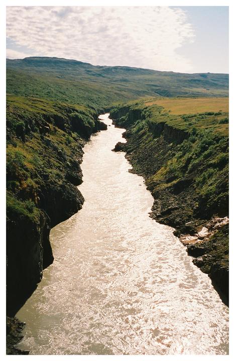 A river flows through a canyon with green hills on either side under a cloudy sky.