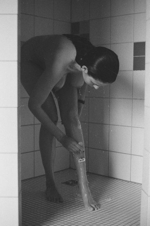 A woman bends over to shave her leg in a tiled shower.