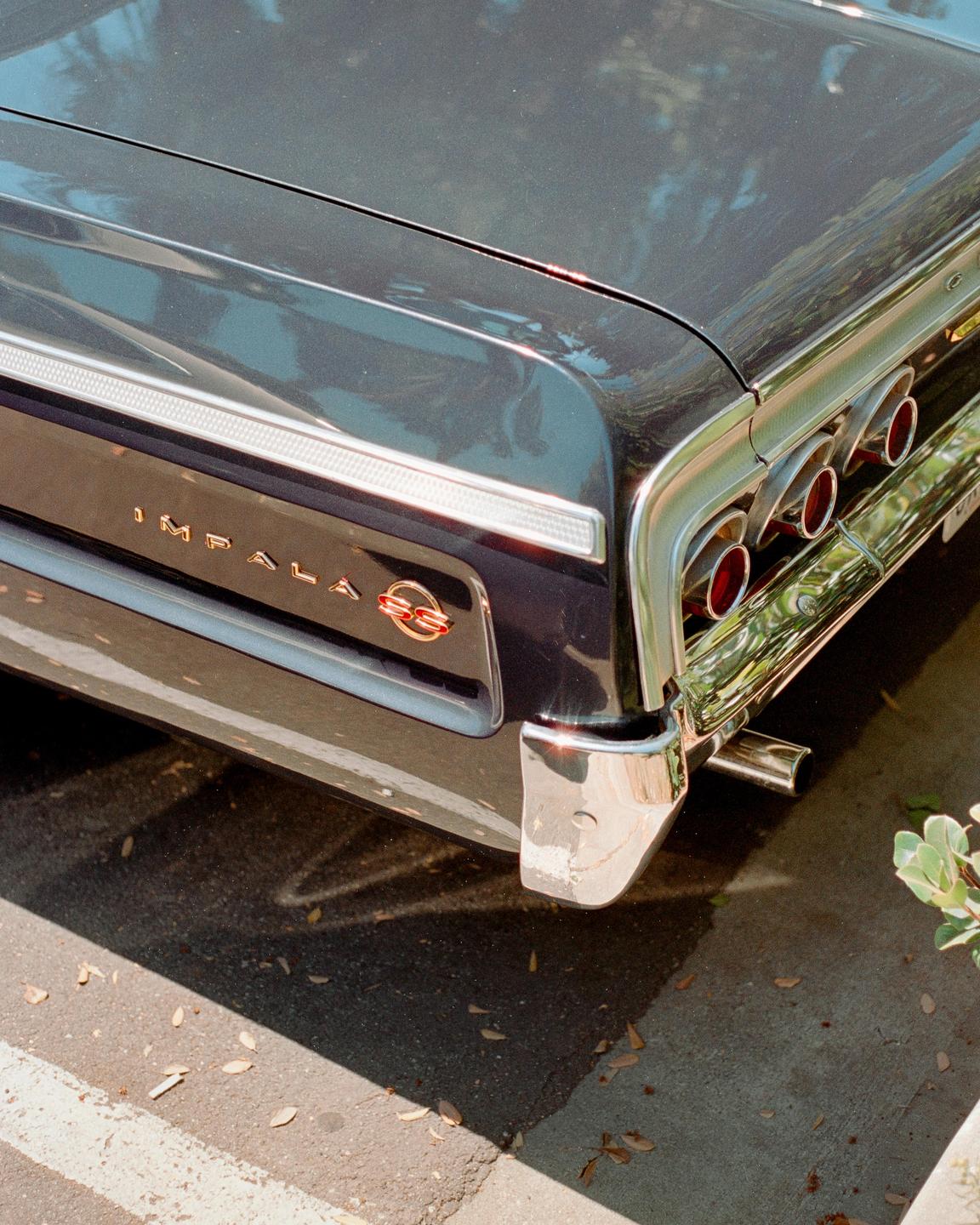 The rear of a dark blue Impala SS is parked on asphalt with fallen leaves.