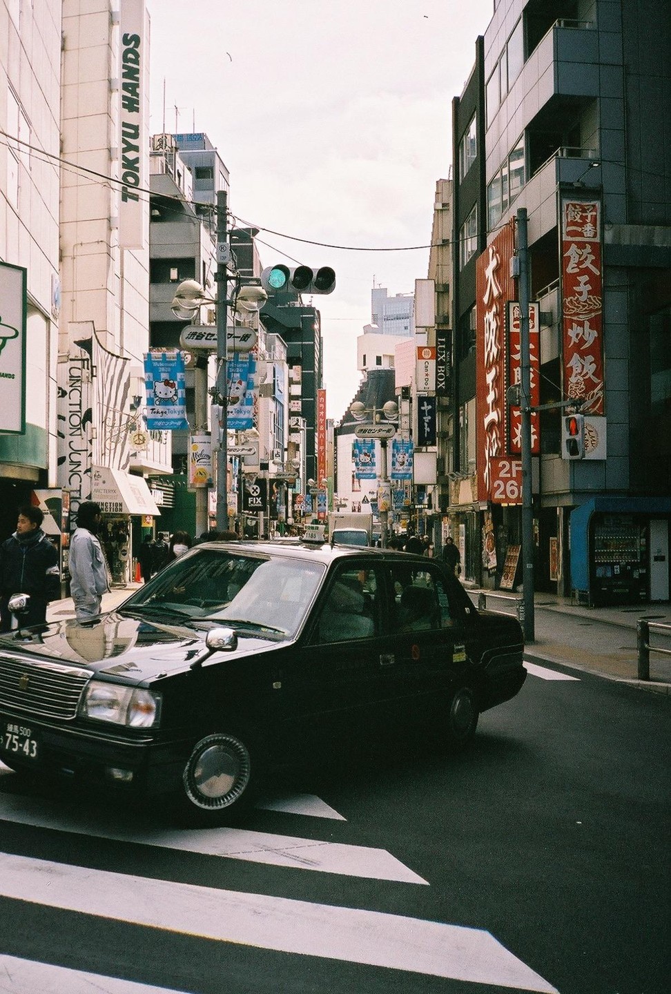 A black taxi drives across a crosswalk on a busy street lined with buildings and signs.