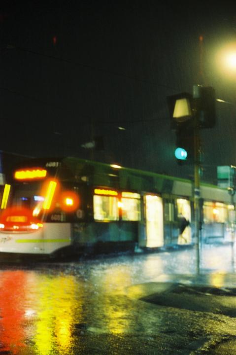 A tram with illuminated windows is stopped at a street corner at night, with reflections of its lights on the wet ground.