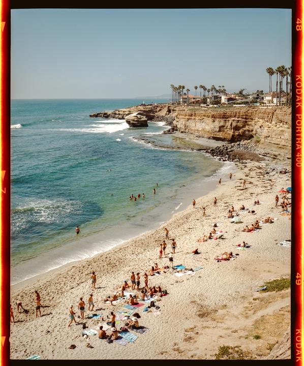 A sandy beach with many people swimming and sunbathing is visible next to a cliff with houses and palm trees.