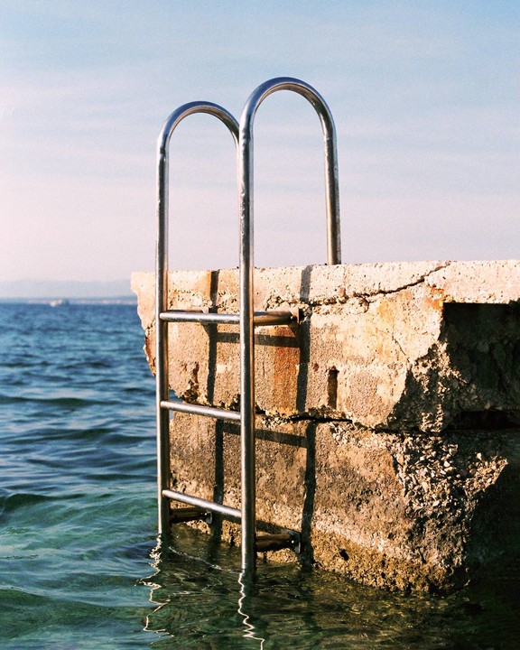 A metal ladder extends from a concrete pier into the clear blue sea under a bright sky.