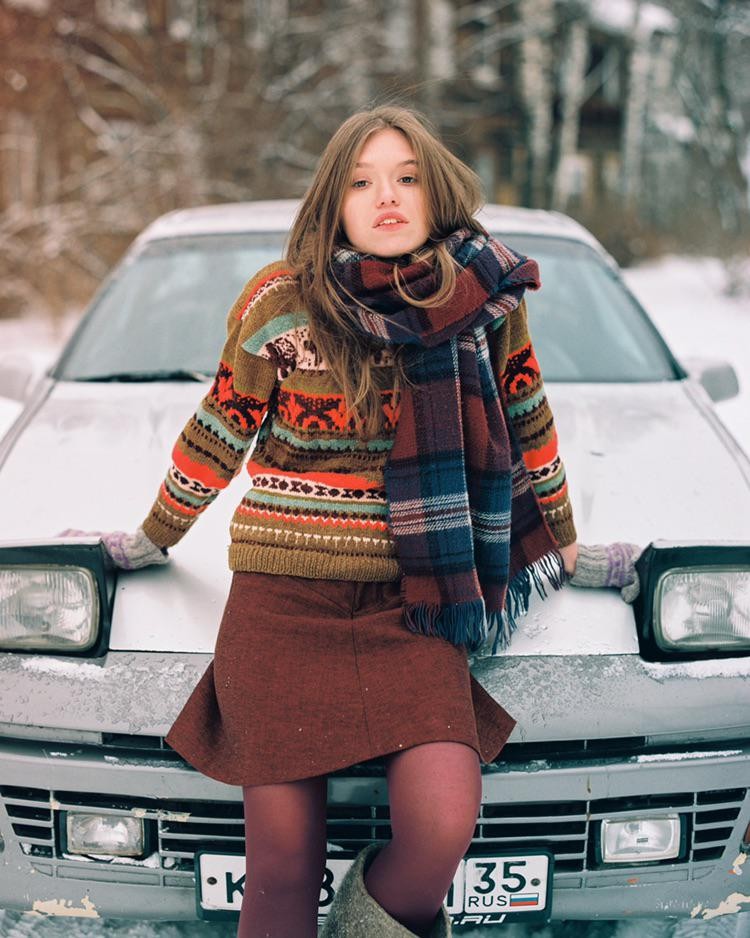 A woman wearing a colorful sweater and scarf leans on the hood of a car in a snowy landscape.