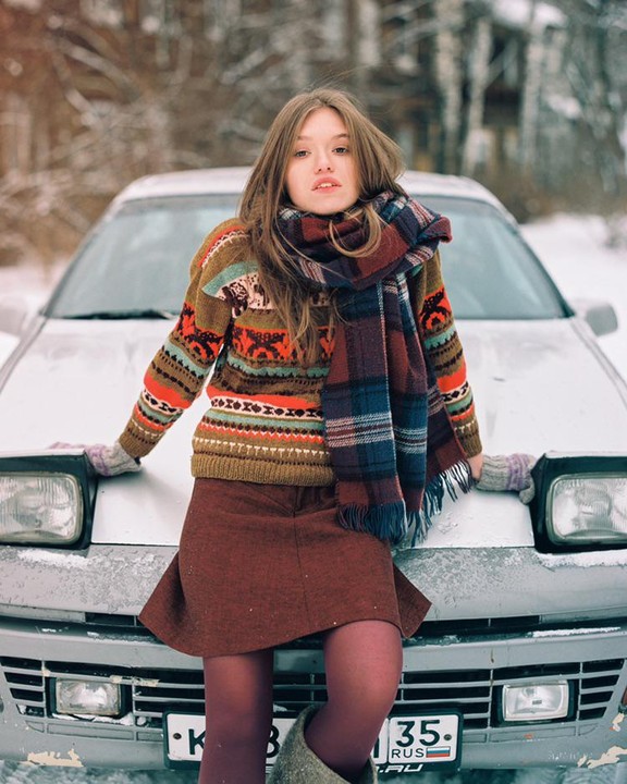 A woman wearing a colorful sweater and scarf leans on the hood of a car in a snowy landscape.
