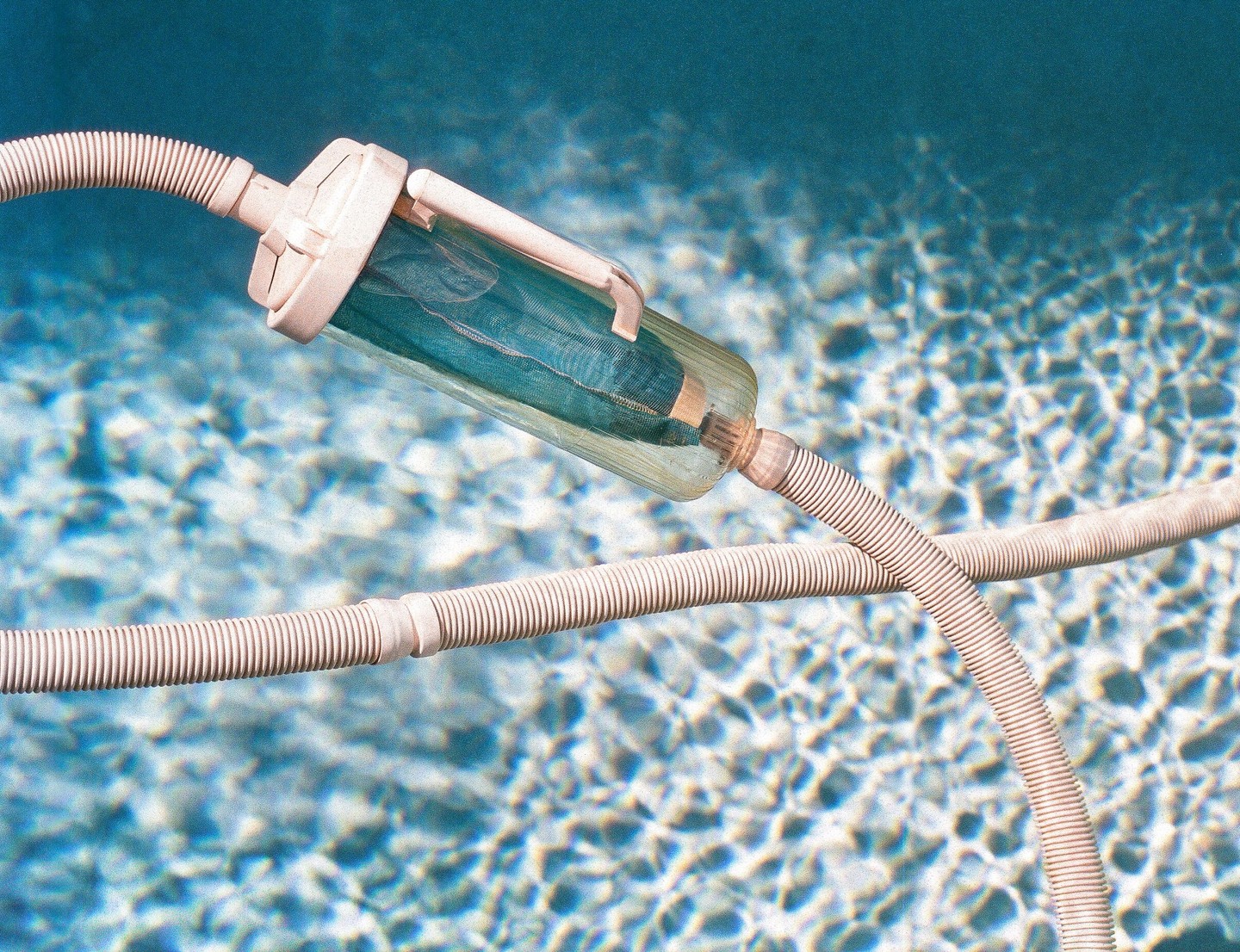 A pool vacuum hose and filter are submerged in clear blue water with sunlight reflecting on the surface.