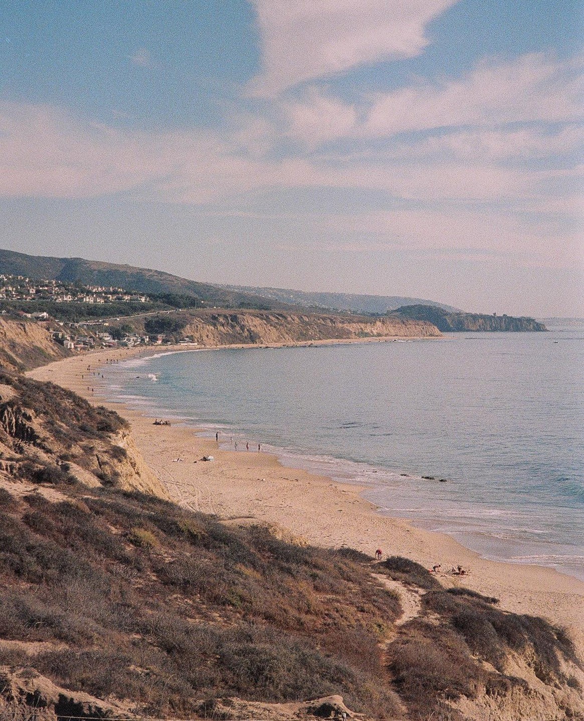 A long stretch of sandy beach with people and waves is visible from a high vantage point, with cliffs and hills in the background.