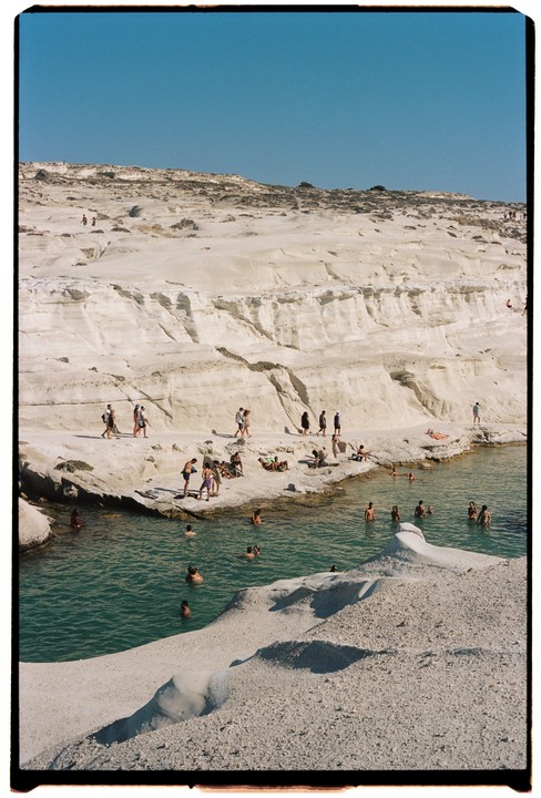 People swim in a cove surrounded by white rock formations under a clear blue sky.