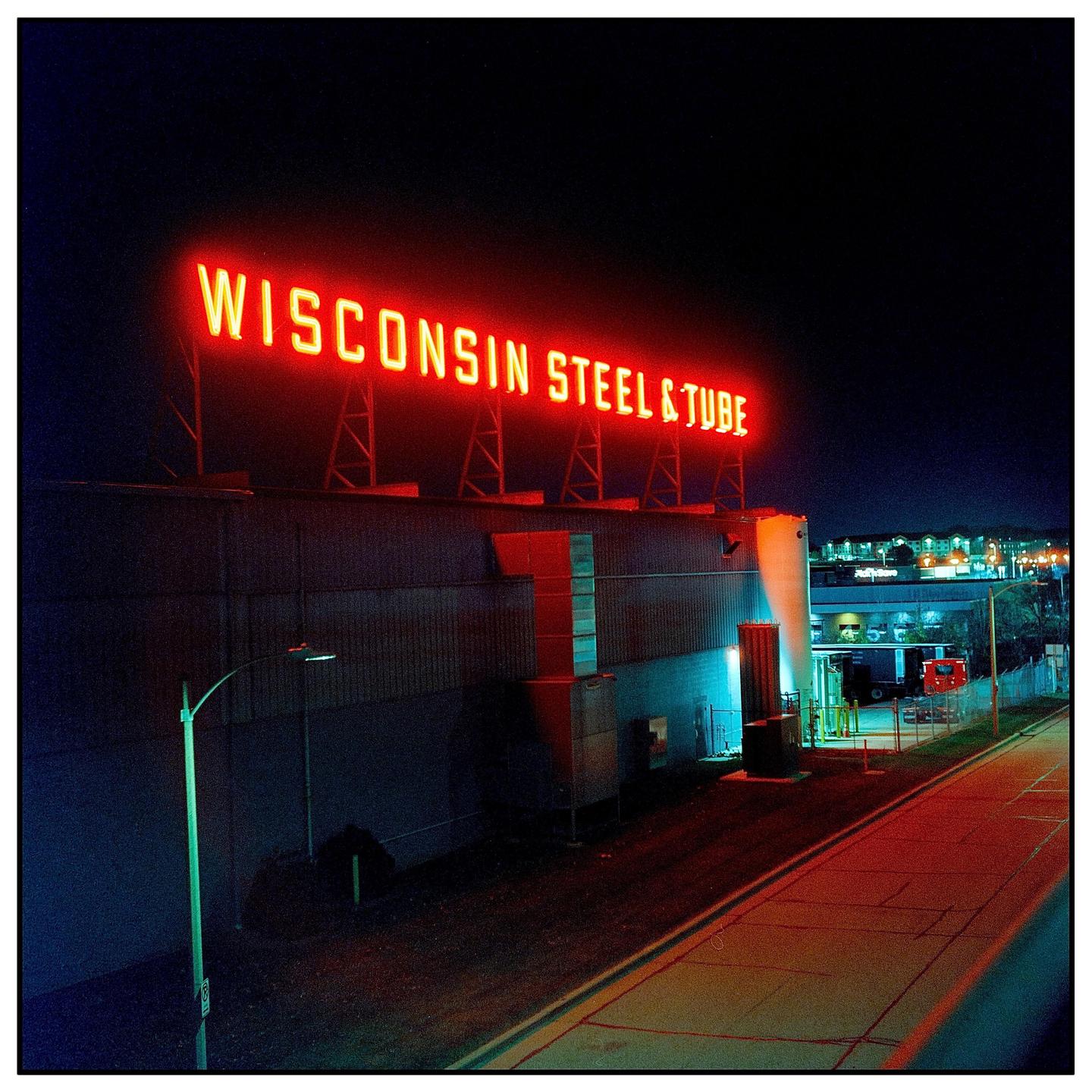 A red neon sign for Wisconsin Steel & Tube illuminates a building at night.