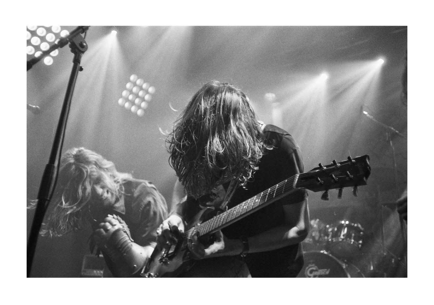 A man with long hair plays an electric guitar on a stage with another musician in the background.