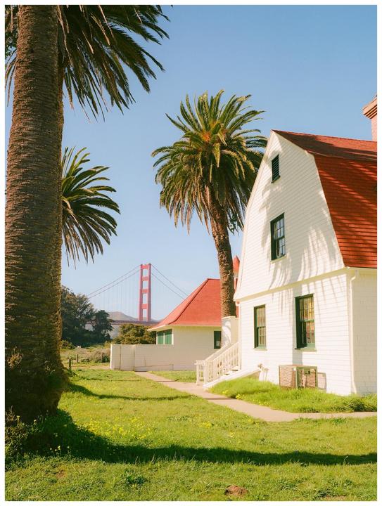 A white house with a red roof stands next to palm trees, with the Golden Gate Bridge visible in the distance.