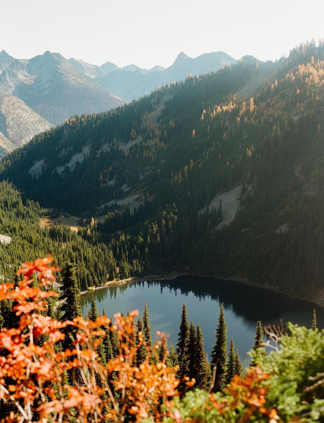 A mountain lake is surrounded by pine trees and autumn foliage under a clear sky.