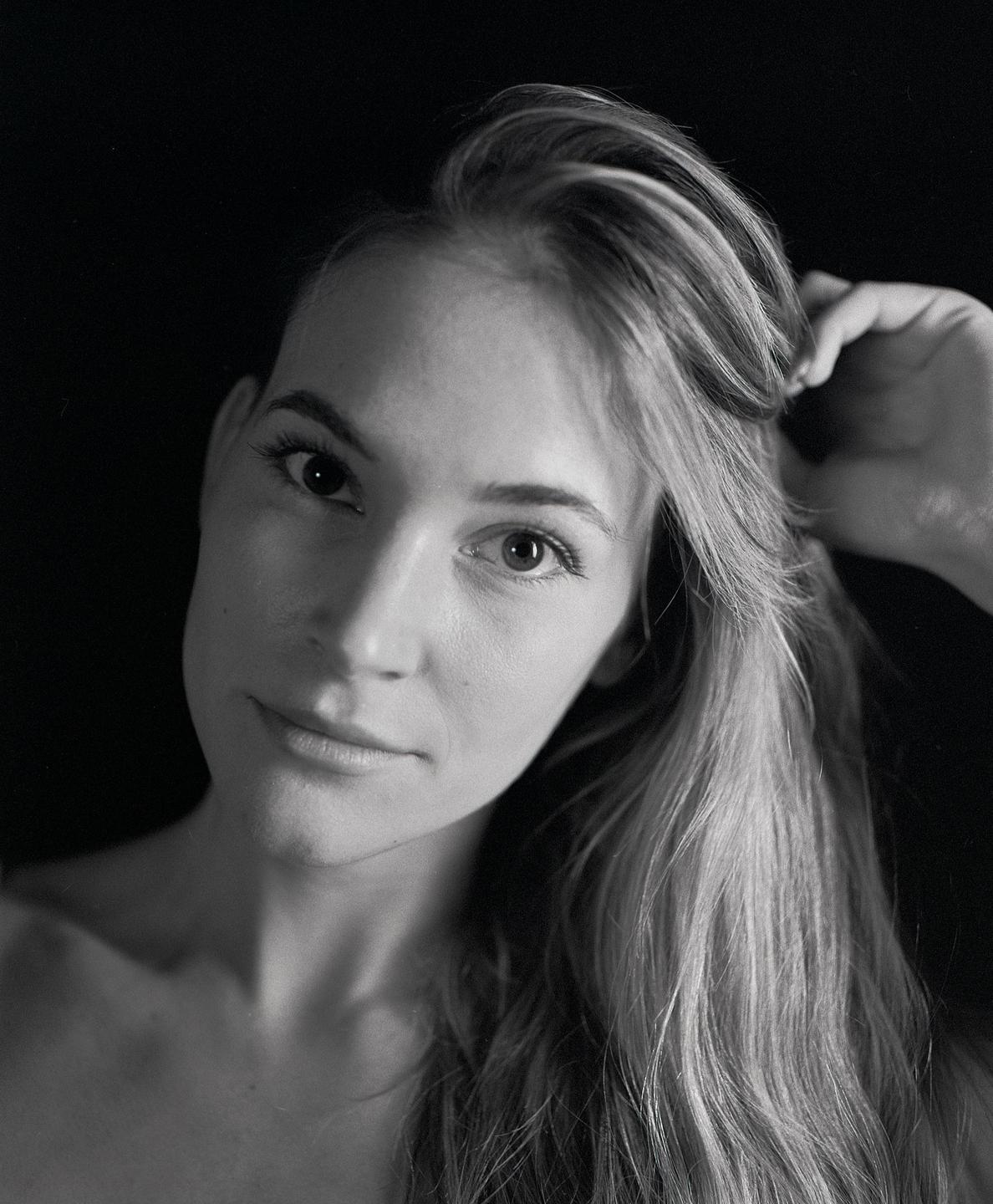 A woman with long hair looks directly at the viewer, touching her hair with one hand.