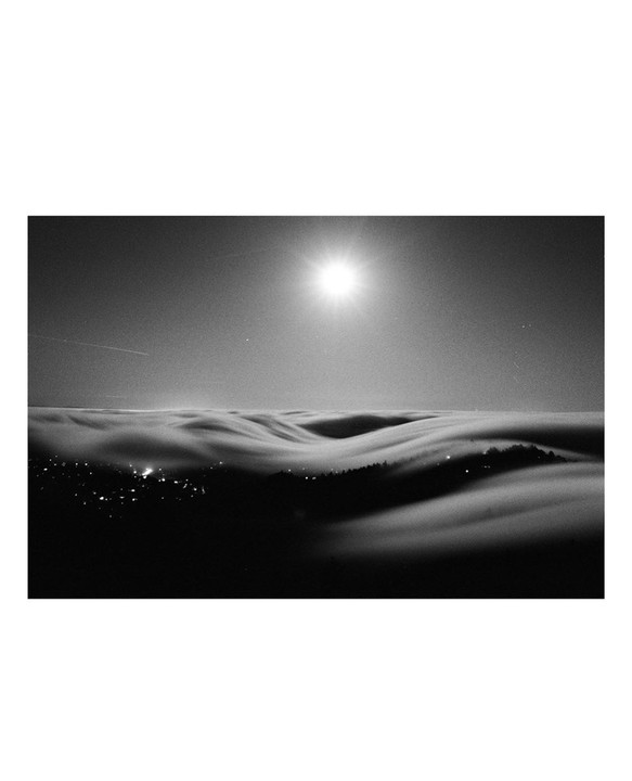 The moon shines brightly above a layer of fog covering a landscape with city lights and trees.