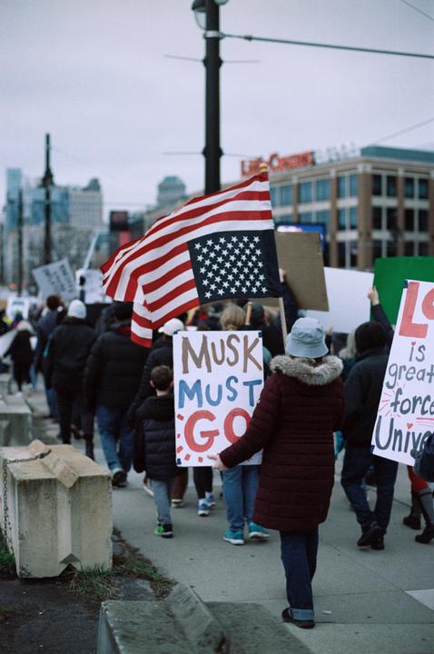 A crowd of people marches down a city street, some holding protest signs and an American flag.