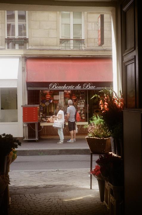 A couple stands in front of a butcher shop with a red awning, viewed from a doorway with plants in the foreground.