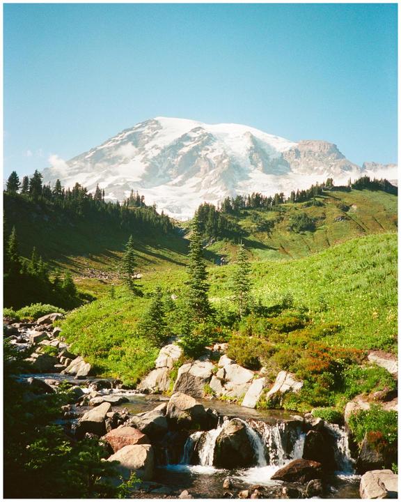 A snow-capped mountain rises above a green valley with a rocky stream and evergreen trees.