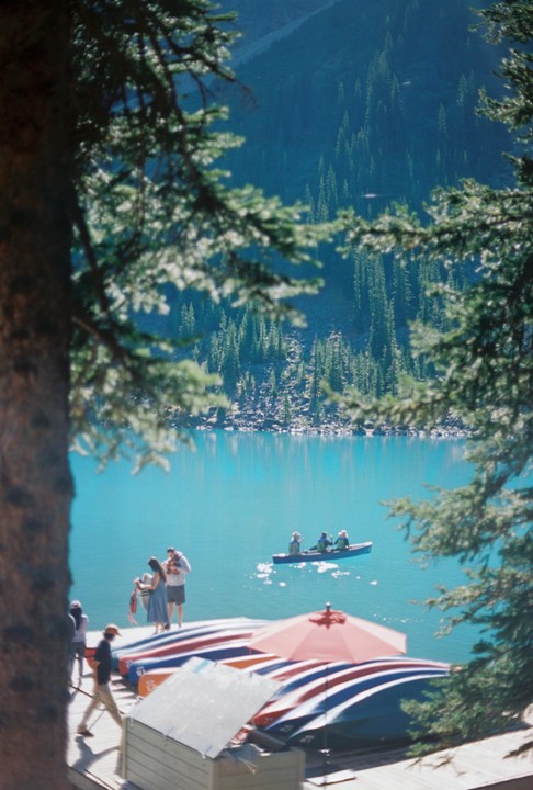 A group of people are on a dock next to a turquoise lake with a canoe and mountains in the background.